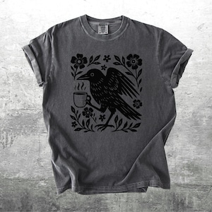 May include: Dark gray t-shirt featuring a black graphic of a crow holding a steaming mug, surrounded by flowers and foliage. The design is centered on the shirt. The shirt has rolled-up sleeves and a crew neck.