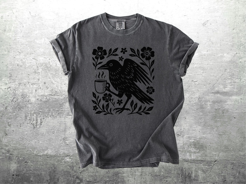 May include: Dark gray t-shirt featuring a black graphic of a crow holding a steaming mug, surrounded by flowers and foliage. The design is centered on the shirt. The shirt has rolled-up sleeves and a crew neck.