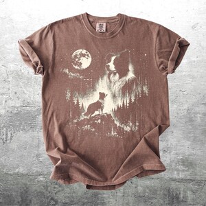 Pu&ograve; includere: T-shirt marrone con una grafica di un cane in una foresta sotto la luna piena. Il design include la silhouette di un cane su una collina, il volto di un cane pi&ugrave; grande e alberi. La maglietta ha un aspetto vintage.