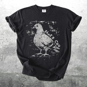 May include: Black t-shirt featuring a distressed white graphic of a pigeon with the text "that's Coo". The shirt has rolled-up sleeves and a crew neck. The design is centered on the front of the shirt.