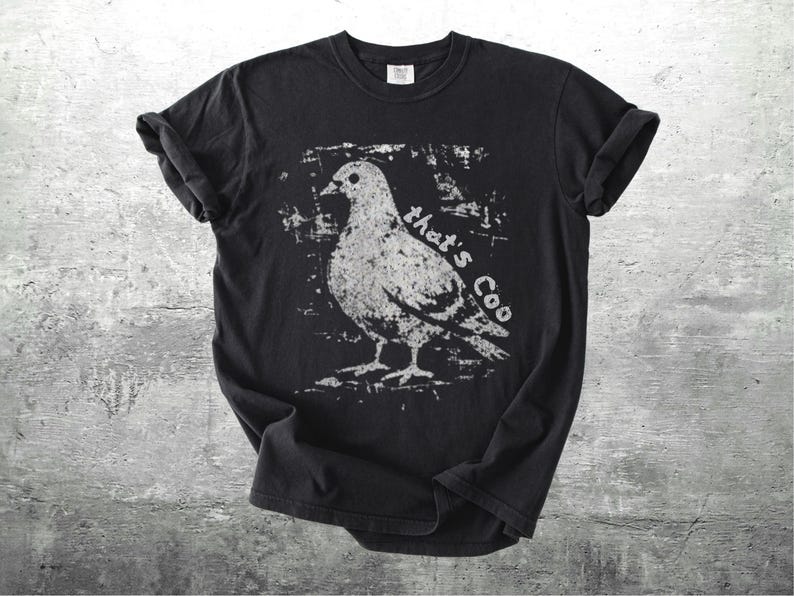May include: Black t-shirt featuring a distressed white graphic of a pigeon with the text "that's Coo". The shirt has rolled-up sleeves and a crew neck. The design is centered on the front of the shirt.