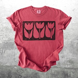 May include: Red t-shirt with a black graphic of three stylized tulips in a rectangular frame. The shirt has rolled-up sleeves and a crew neck. The fabric has a slightly faded appearance.