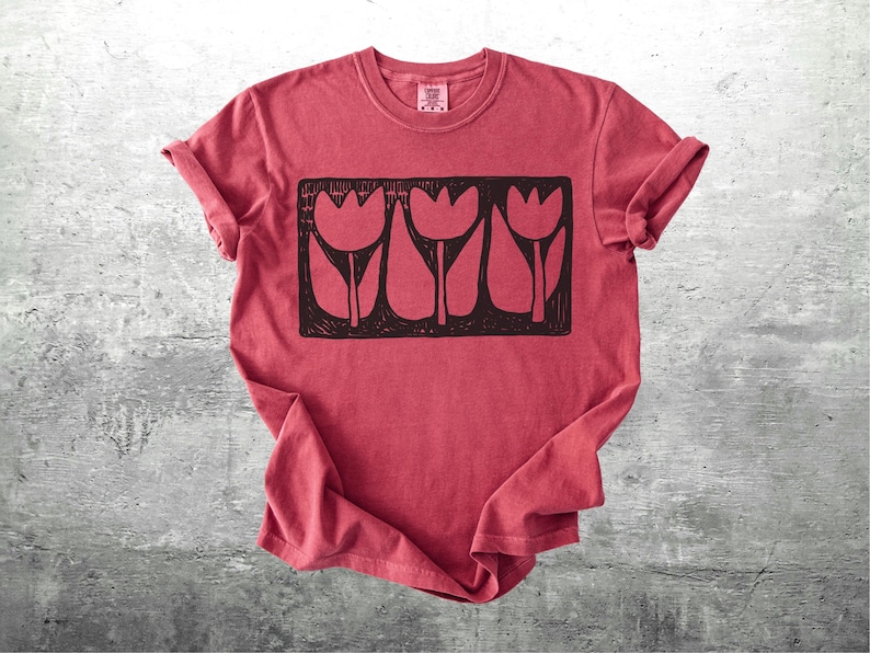 May include: Red t-shirt with a black graphic of three stylized tulips in a rectangular frame. The shirt has rolled-up sleeves and a crew neck. The fabric has a slightly faded appearance.