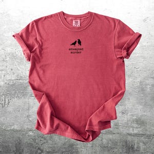 May include: A red t-shirt with rolled-up sleeves. The shirt features a small graphic of two black birds above the words "attempted murder" in a simple font. The shirt is displayed on a gray, textured surface.