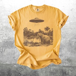 May include: Mustard yellow t-shirt featuring a brown graphic of a capybara standing near a body of water, with a UFO hovering above. The design includes palm trees and other foliage. The shirt has a crew neck and short sleeves.