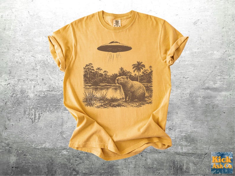 May include: Mustard yellow t-shirt featuring a brown graphic of a capybara standing near a body of water, with a UFO hovering above. The design includes palm trees and other foliage. The shirt has a crew neck and short sleeves.