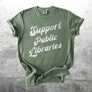 May include: Olive green t-shirt with rolled sleeves. The shirt features the text "Support Public Libraries" in a distressed white font. The shirt is made of a soft material and is suitable for casual wear.