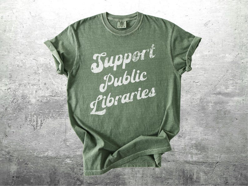May include: Olive green t-shirt with rolled sleeves. The shirt features the text "Support Public Libraries" in a distressed white font. The shirt is made of a soft material and is suitable for casual wear.
