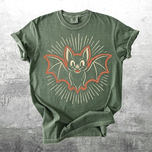 May include: An olive green short-sleeve t-shirt featuring a central graphic of a smiling bat with orange outlines and cream-colored radiating lines. The shirt has a crew neckline and a vintage-style wash, with the brand name Kick Ash Co. visible.