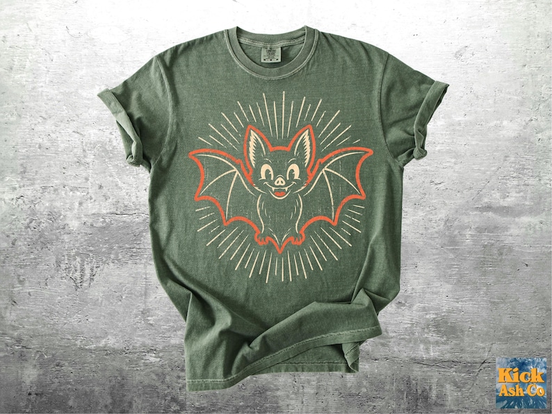 May include: An olive green short-sleeve t-shirt featuring a central graphic of a smiling bat with orange outlines and cream-colored radiating lines. The shirt has a crew neckline and a vintage-style wash, with the brand name Kick Ash Co. visible.