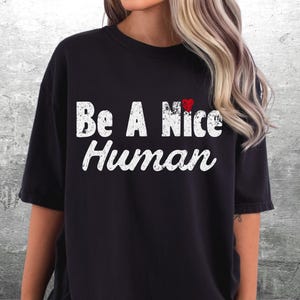 K&ouml;nnte beinhalten: Schwarzes T-Shirt mit dem wei&szlig;en, verwitterten Schriftzug "Be A Nice Human". Ein kleines rotes Herz befindet sich &uuml;ber dem Wort "Nice". Das Shirt hat einen l&auml;ssigen Stil.