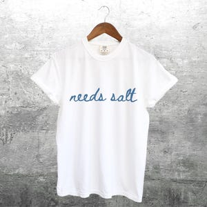 May include: White t-shirt with blue embroidered text that reads "needs salt".