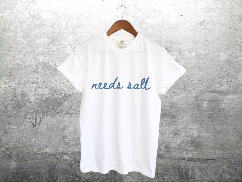 May include: White t-shirt with blue embroidered text that reads "needs salt".