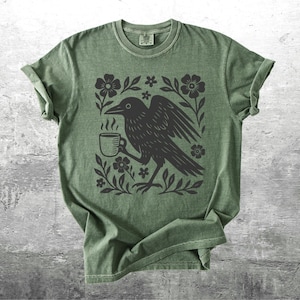 May include: Green t-shirt featuring a black crow holding a coffee cup, surrounded by floral elements. The design is printed on a soft, olive-green tee. The shirt has short sleeves and a crew neck, perfect for casual wear.