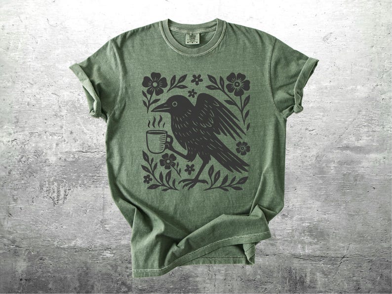 May include: Green t-shirt featuring a black crow holding a coffee cup, surrounded by floral elements. The design is printed on a soft, olive-green tee. The shirt has short sleeves and a crew neck, perfect for casual wear.