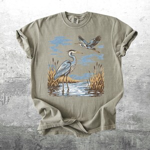 May include: A sage green t-shirt featuring a nature scene with two herons. One heron stands in water, while another flies above. The design includes blue sky, brown reeds, and water reflections, creating a serene, outdoorsy theme.