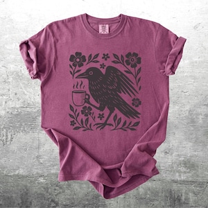 May include: A maroon t-shirt featuring a black illustration of a raven holding a coffee cup, surrounded by floral elements. The design is centered on the shirt, which is folded on a gray surface.