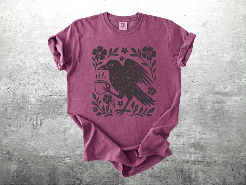 May include: A maroon t-shirt featuring a black illustration of a raven holding a coffee cup, surrounded by floral elements. The design is centered on the shirt, which is folded on a gray surface.