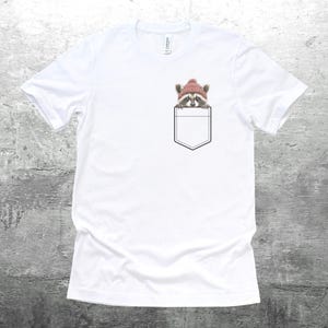 May include: White t-shirt with a pocket featuring a raccoon wearing a pink beanie hat.