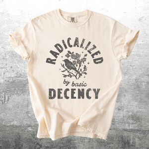 May include: Cream-colored t-shirt with the text "RADICALIZED by basic DECENCY" in a distressed, dark gray font. A bird perched on a floral branch is centered above the words. The shirt is folded.