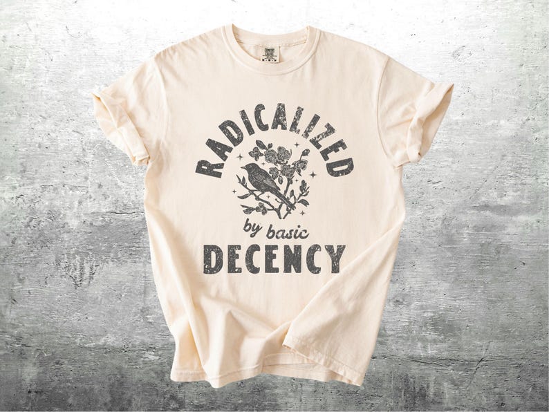 May include: Cream-colored t-shirt with the text "RADICALIZED by basic DECENCY" in a distressed, dark gray font. A bird perched on a floral branch is centered above the words. The shirt is folded.