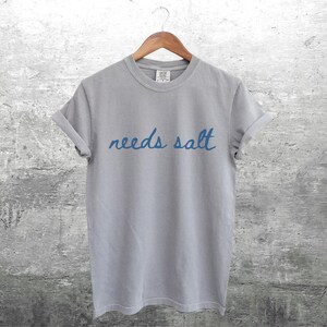 May include: A gray t-shirt with the text "needs salt" written in blue cursive.