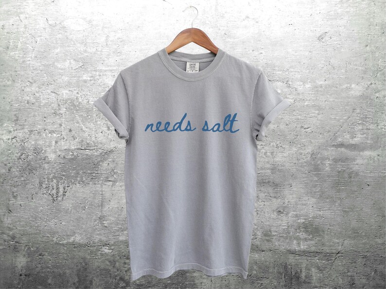 May include: A gray t-shirt with the text "needs salt" written in blue cursive.