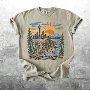 May include: A beige short-sleeve t-shirt featuring a graphic print of a raccoon in the foreground with a Seattle skyline, the Space Needle, a mountain, and a rising sun in the background, framed by two tall green pine trees on either side.