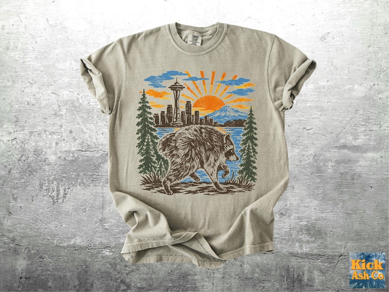 May include: A beige short-sleeve t-shirt featuring a graphic print of a raccoon in the foreground with a Seattle skyline, the Space Needle, a mountain, and a rising sun in the background, framed by two tall green pine trees on either side.