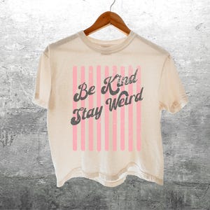 May include: A cream-coloured short-sleeve t-shirt featuring a graphic design with vertical light pink stripes and the dark grey text "Be Kind Stay Weird" in a distressed, retro-inspired font, hanging on a wooden hanger.