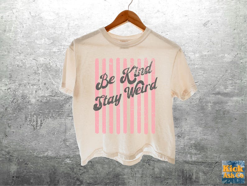 May include: A cream-coloured short-sleeve t-shirt featuring a graphic design with vertical light pink stripes and the dark grey text "Be Kind Stay Weird" in a distressed, retro-inspired font, hanging on a wooden hanger.
