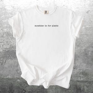 May include: White t-shirt with the text "sunshine is for plants" in a simple, black font. The shirt is made of a soft material and has a classic crew neck design. The shirt is displayed on a grey, textured surface.