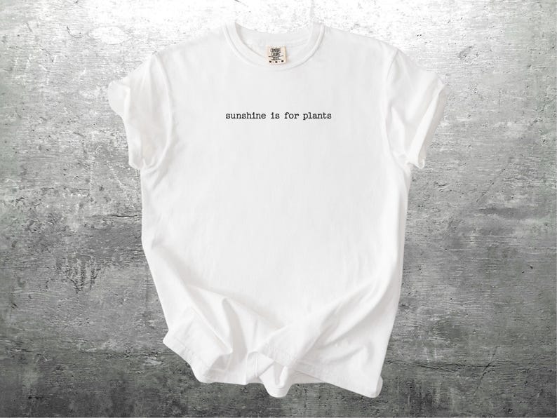 May include: White t-shirt with the text "sunshine is for plants" in a simple, black font. The shirt is made of a soft material and has a classic crew neck design. The shirt is displayed on a grey, textured surface.