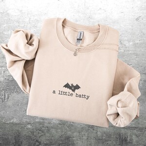 May include: A beige sweatshirt with the text "a little batty" and a bat graphic. A silver necklace with a small charm is on the sweatshirt. The sleeves are folded. The sweatshirt is on a gray surface.