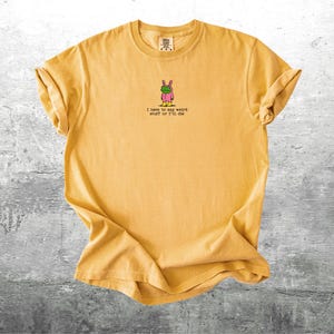 May include: Mustard yellow t-shirt with rolled sleeves. The shirt features a small graphic of a cartoon bunny wearing a green mask and pink outfit. Below the graphic is the text: "I have to say weird stuff or I'll die."