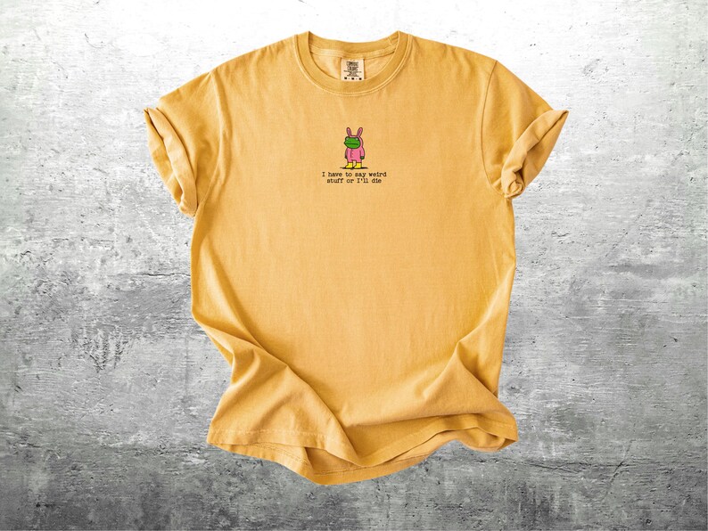 May include: Mustard yellow t-shirt with rolled sleeves. The shirt features a small graphic of a cartoon bunny wearing a green mask and pink outfit. Below the graphic is the text: "I have to say weird stuff or I'll die."
