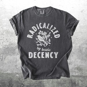 May include: Dark gray t-shirt with rolled sleeves. The shirt features the text "RADICALIZED by basic DECENCY" in white, with a bird and floral graphic. The shirt has a vintage, worn look.