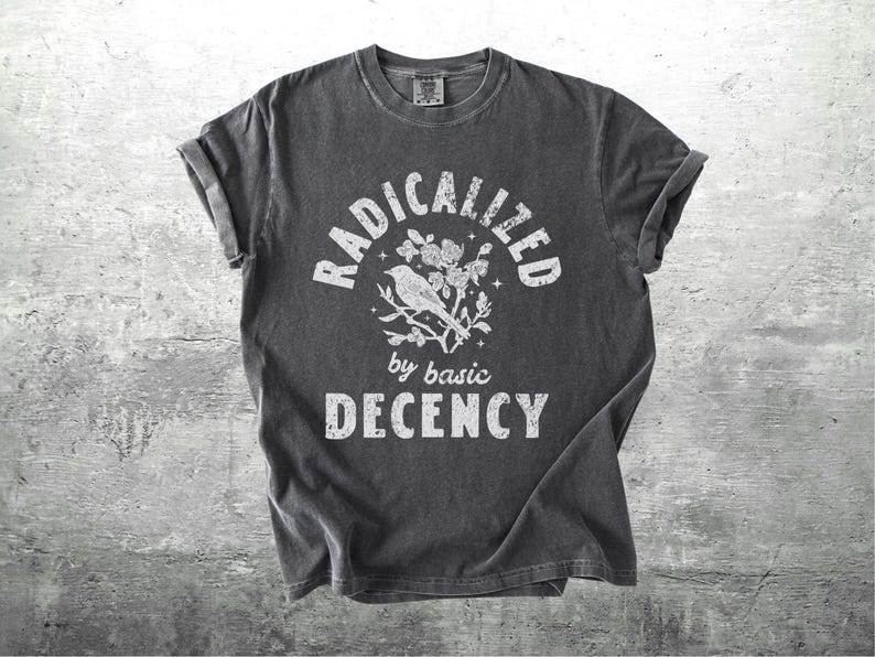 May include: Dark gray t-shirt with rolled sleeves. The shirt features the text "RADICALIZED by basic DECENCY" in white, with a bird and floral graphic. The shirt has a vintage, worn look.
