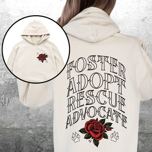 Pu&ograve; includere: Una felpa con cappuccio color crema con una grafica in bianco e nero sul retro che dice "Foster Adopt Rescue Advocate" con una rosa rossa e impronte di zampe. La parte anteriore della felpa con cappuccio ha una piccola grafica di rosa rossa.