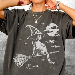 May include: A charcoal gray short-sleeve t-shirt featuring a distressed white graphic print of a cat wearing a pointed witch hat, sitting on a broomstick against a full moon and clouds. The bottom right corner displays the text Kick Ash-Co.