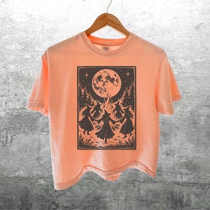 May include: A peach-colored short-sleeve t-shirt featuring a dark brown rectangular graphic print on the front. The illustration depicts four figures in pointed hats dancing in a circle around a bonfire under a large full moon and pine trees.