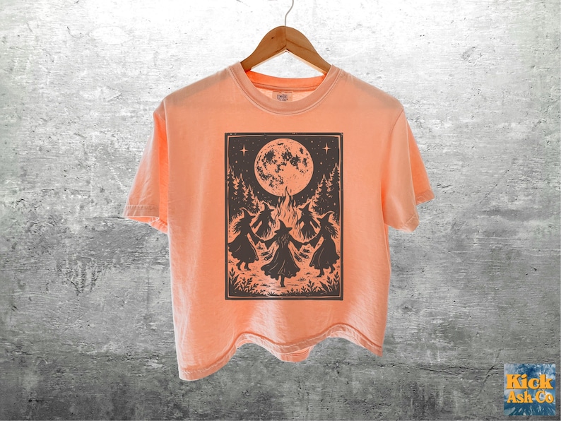 May include: A peach-colored short-sleeve t-shirt featuring a dark brown rectangular graphic print on the front. The illustration depicts four figures in pointed hats dancing in a circle around a bonfire under a large full moon and pine trees.