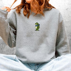 Puede incluir: Una sudadera gris con un peque&ntilde;o bordado de una rana con una bandana azul. La rana es verde con manchas amarillas. La sudadera tiene mangas largas y cuello redondo. La persona lleva vaqueros azules.