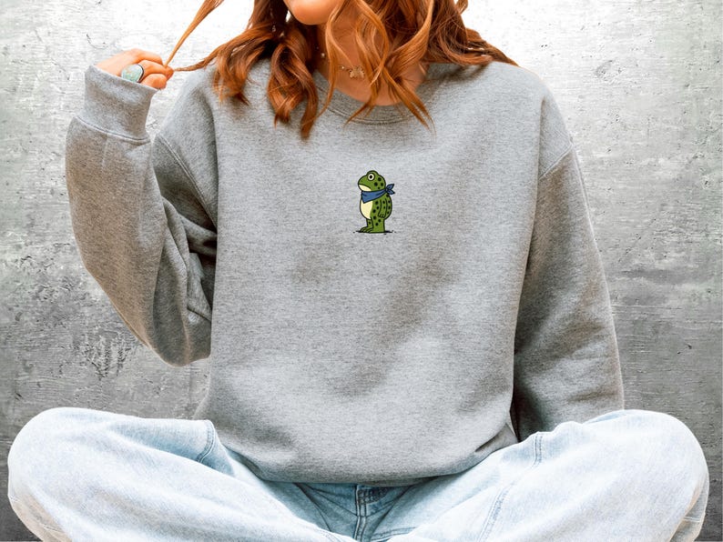Puede incluir: Una sudadera gris con un peque&ntilde;o bordado de una rana con una bandana azul. La rana es verde con manchas amarillas. La sudadera tiene mangas largas y cuello redondo. La persona lleva vaqueros azules.