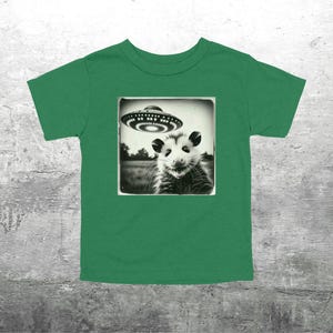 May include: Green t-shirt with a black and white image of an opossum and a UFO. The opossum is in a square frame. The UFO is above the opossum. The shirt is made of a soft material. The design is detailed.