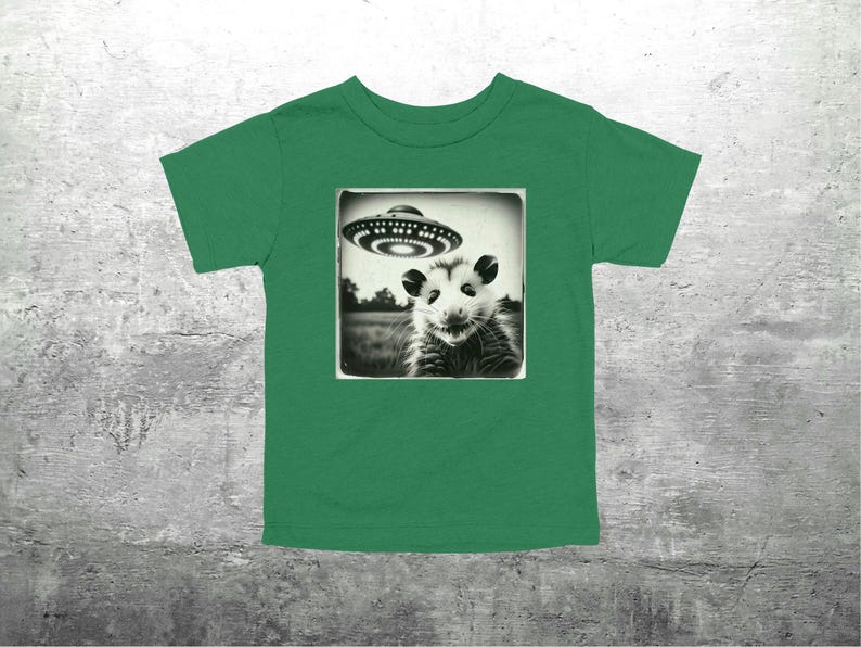 May include: Green t-shirt with a black and white image of an opossum and a UFO. The opossum is in a square frame. The UFO is above the opossum. The shirt is made of a soft material. The design is detailed.