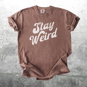 K&ouml;nnte beinhalten: Braunes T-Shirt mit dem wei&szlig;en Schriftzug "Stay Weird". Das Shirt hat einen Rundhalsausschnitt und hochgekrempelte &Auml;rmel. Der Stoff hat einen leicht verblichenen, Vintage-Look.