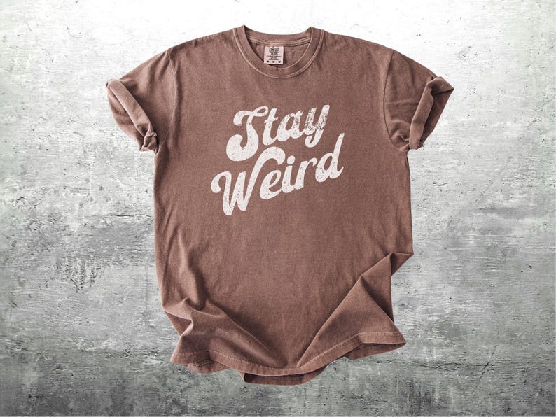 K&ouml;nnte beinhalten: Braunes T-Shirt mit dem wei&szlig;en Schriftzug "Stay Weird". Das Shirt hat einen Rundhalsausschnitt und hochgekrempelte &Auml;rmel. Der Stoff hat einen leicht verblichenen, Vintage-Look.