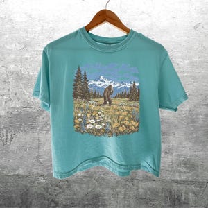 Puede incluir: Una camiseta de color turquesa con un gr&aacute;fico de un Bigfoot en un campo con monta&ntilde;as y &aacute;rboles. El Bigfoot es marr&oacute;n y las monta&ntilde;as son azules y blancas. La camiseta est&aacute; colgada de una percha de madera.