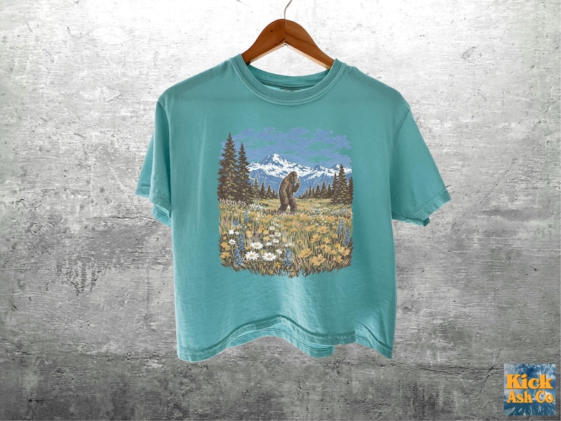 Puede incluir: Una camiseta de color turquesa con un gr&aacute;fico de un Bigfoot en un campo con monta&ntilde;as y &aacute;rboles. El Bigfoot es marr&oacute;n y las monta&ntilde;as son azules y blancas. La camiseta est&aacute; colgada de una percha de madera.